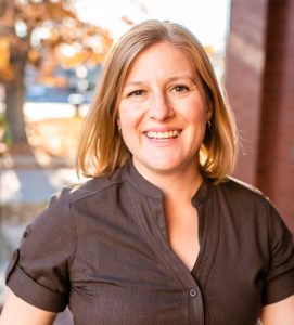 Hannah Curtis, LCSW is seen smiling, wearing a brown dress with autumn tree in the background