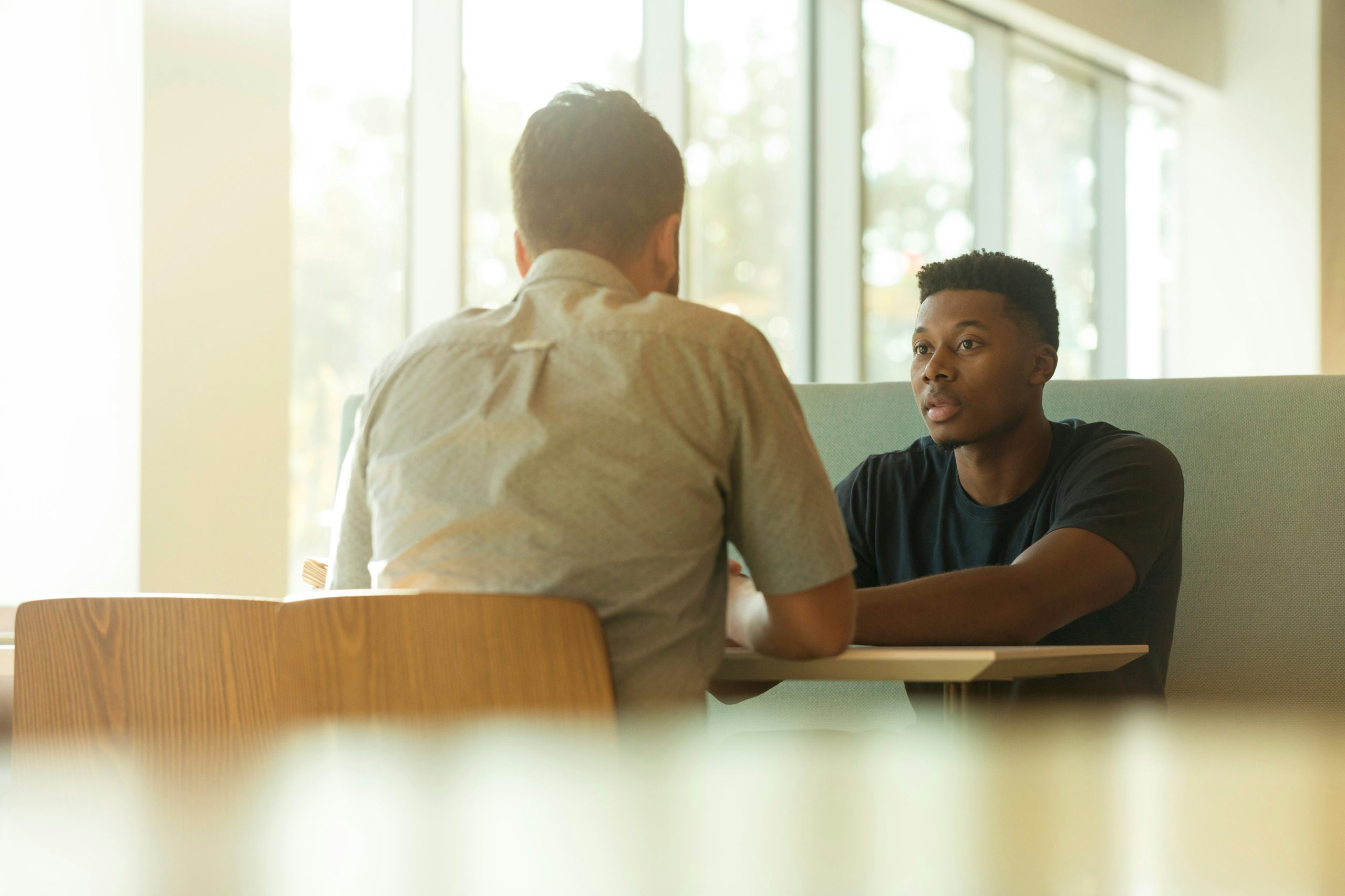 Two men sit together. Only one of their faces can be seen. They are in conversation.